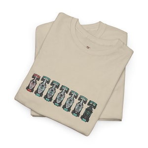 Puede incluir: Dos camisetas beige dobladas y apiladas. La camiseta superior presenta un gráfico colorido de siete coches de carreras en fila. Los coches tienen varios esquemas de color, incluyendo rojo, turquesa y negro.