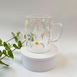 May include: A clear glass mug with a handle, decorated with a colourful floral pattern. The mug is sitting on a white pedestal.