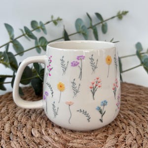 May include: A cream-coloured ceramic mug adorned with colourful floral illustrations. The flowers include pink, yellow, purple, and blue blooms, with green foliage. The mug has a curved handle and a wide opening, suitable for hot drinks.