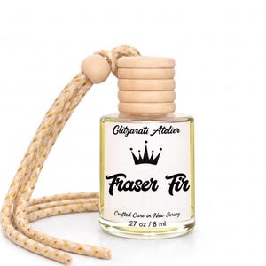 May include: A small clear glass bottle filled with yellow liquid, featuring a light wood cap and a beige hanging cord. The label reads Glitzarati Atelier, a crown icon, Fraser Fir, Crafted Care in New Jersey, and .27 oz / 8 ml.