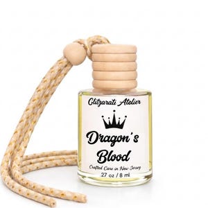 May include: A small clear glass bottle with a light yellow liquid, a ribbed wooden cap, and a beige braided hanging cord. The white label features a black crown icon and the text Glitzarati Atelier, Dragon's Blood, Crafted Care in New Jersey, .27 oz / 8 ml.