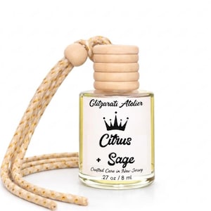 May include: A small clear glass car air freshener bottle with a light yellow liquid, a ribbed wooden cap, and a beige braided hanging cord. The white label features a black crown icon and the text Glitzarati Atelier Citrus + Sage .27 oz / 8 ml.