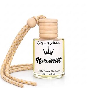 May include: A clear glass car air freshener bottle with a light wooden cap and a beige braided hanging cord. The white label features a black crown icon, the text Glitzarati Atelier, Narcissist, Crafted Care in New Jersey, and .27 oz / 8 ml.