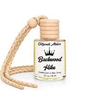 May include: A small clear glass bottle containing liquid with a light wooden cap and a braided beige hanging cord. The white label features the text Glitzarati Atelier, a crown icon, Backwood Hike, Crafted Core in New Jersey, and .27 oz / 8 ml.