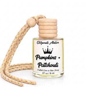 May include: A small clear glass bottle filled with yellow liquid, featuring a light wood cap and a beige braided hanging cord. The label reads Glitzarati Atelier, Pumpkins + Patchouli, Crafted Care in New Jersey, and .27 oz / 8 ml.