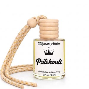 May include: A small clear glass bottle filled with light yellow liquid, featuring a wooden cap and a beige hanging cord. The label reads Glitzarati Atelier, a crown icon, Patchouli, Crafted Care in New Jersey, and .27 oz / 8 ml.