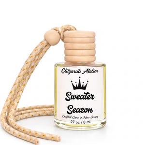 May include: A clear glass bottle with a wooden cap and a beige rope. The label reads "Sweater Season" with a crown graphic. The bottle contains 27 oz / 8 ml of liquid. The text "Glitzarati Atelier" is also visible.