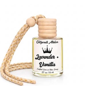 May include: A clear glass car air freshener bottle with a light yellow liquid, a ribbed wooden cap, and a beige braided hanging cord. The white label features a black crown icon and the text Glitzarati Atelier, Lavender + Vanilla, Crafted Care in New Jersey, .27 oz / 8 ml.