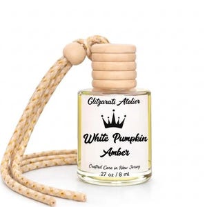 May include: A small glass car air freshener bottle with a light wooden cap and a beige braided hanging cord. The white label features a black crown icon and the text Glitzarati Atelier, White Pumpkin Amber, Crafted Care in New Jersey, .27 oz / 8 ml.