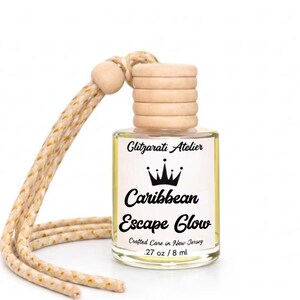 May include: A small glass bottle of fragrance with a light wooden cap and a beige cord featuring gold flecks. The white label displays the text Glitzarati Atelier, a crown icon, Caribbean Escape Glow, Crafted Care in New Jersey, and .27 oz / 8 ml.