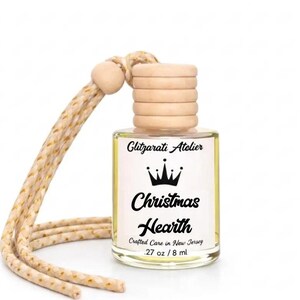 May include: A small clear glass bottle with a light yellow liquid, featuring a wooden cap and a braided beige hanging cord. The label reads Glitzarati Atelier, a crown icon, Christmas Hearth, Crafted Care in New Jersey, and .27 oz / 8 ml.