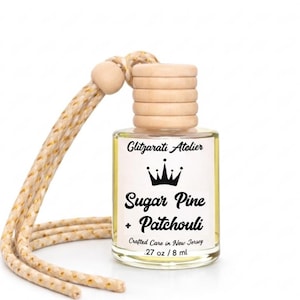 May include: A small, clear glass bottle with a wooden cap and a cream-colored cord. The label reads "Glitzarati Atelier" with "Sugar Pine + Patchouli" below a crown graphic. The bottle contains 0.27 oz / 8 ml of liquid.
