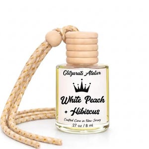 May include: A clear glass car air freshener bottle with a light wooden cap and a beige braided hanging cord. The label features the text Glitzarati Atelier, a crown icon, White Peach + Hibiscus, Crafted Care in New Jersey, and .27 oz / 8 ml.