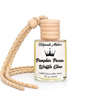 May include: A small clear glass bottle with a light wooden cap and a beige braided cord. The label reads Glitzarati Atelier, a crown icon, Pumpkin Pecan Waffle Glow, Crafted Care in New Jersey, and .27 oz / 8 ml. The bottle contains liquid.