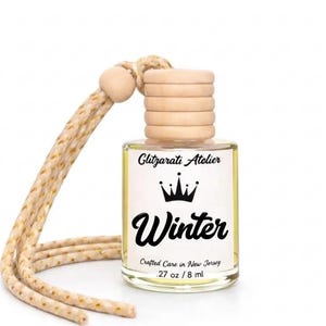 May include: A clear glass car air freshener bottle with a light yellow liquid, featuring a wooden cap and a beige braided hanging cord. The label reads Glitzarati Atelier, a crown icon, Winter, Crafted Care in New Jersey, and .27 oz / 8 ml.