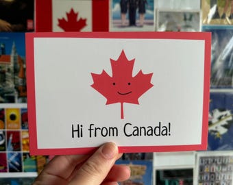 May include: A rectangular postcard with a red border and a white interior. The postcard features a red maple leaf with a smiling face and the text "Hi from Canada!"