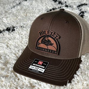 Könnte beinhalten: Braune und beigefarbene Trucker-Kappe mit einem braunen Leder-Patch, auf dem in weißen Buchstaben "Built Snorkeler" steht. Die Kappe hat einen schwarzen verstellbaren Riemen mit einem weißen Etikett, auf dem "Richardson 112 Original Trucker Adjustable" steht.