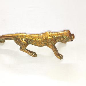 Könnte beinhalten: Eine goldfarbene Metallfigur eines Leoparden in einer laufenden Pose. Die Figur ist mit Flecken verziert und hat ein realistisches Aussehen.
