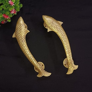 May include: Two brass-coloured door handles shaped like fish. The handles have a textured scale pattern and are curved. The fish-shaped handles are on a black background. Decorative hardware.