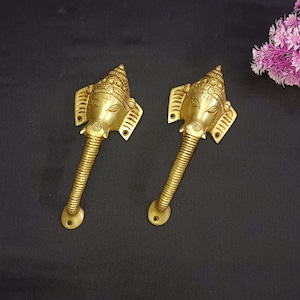 May include: Two gold-toned door handles shaped like the head of an elephant god with intricate details. The handles are on a black background.