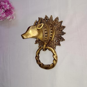 May include: A brass door knocker featuring a stylized boar's head with a ring handle. The boar's head is detailed with etched lines and a sunburst-like design around it. The knocker is a warm gold color.