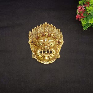 Puede incluir: Una máscara de metal de color dorado con una expresión feroz. La máscara tiene un diseño detallado con tallas intrincadas y una superficie texturizada en relieve.