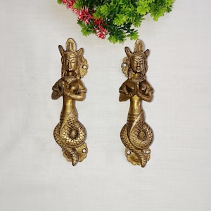 May include: Two gold-toned door handles shaped like a woman with a snake tail. The woman is in a prayer position with her hands clasped together. The handles are ornate and detailed.