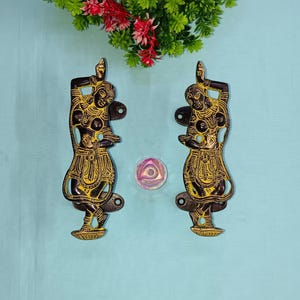 Könnte beinhalten: Zwei goldfarbene Metall-Türgriffe in Form von tanzenden Figuren. Die Figuren sind in einer schwarzen Silhouette mit goldenen Akzenten. Die Griffe befinden sich auf einem hellblauen Hintergrund.
