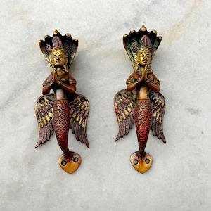 May include: Two ornate brass door handles shaped like a mermaid with wings. The mermaids are in a prayer position with their hands clasped together. The handles are painted in shades of gold, red, and green.