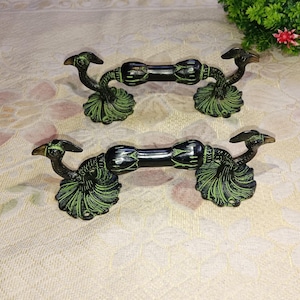 Puede incluir: Dos tiradores de puerta con forma de pavo real con acabado negro y detalles verdes. Los tiradores son de metal y tienen un diseño decorativo.