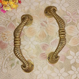 Peut inclure: Deux poignées de porte en laiton ornées d'un motif de feuilles. Les poignées sont courbées et ont un anneau décoratif autour de la base.