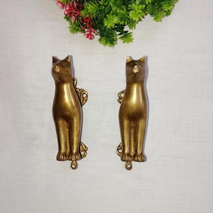 May include: Two brass cat-shaped door knockers. The knockers are golden-toned, with detailed features including ears, eyes, and paws. Each cat has an ornate backplate and a small ring at the bottom. The background is a white surface with green and red decorative foliage.
