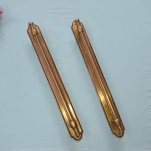 Peut inclure: Deux poignées de meuble antiques de couleur bronze. Chaque poignée est rectangulaire avec un motif central rainuré et des embouts décoratifs. Les poignées mesurent environ 30 cm de long, sur fond bleu clair.
