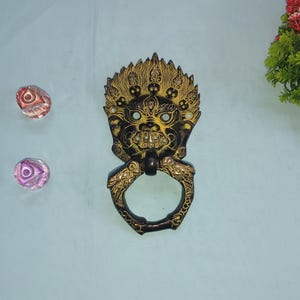 Peut inclure: Un heurtoir de porte de style antique orné, de couleur bronze foncé, avec un motif de visage détaillé et une poignée en anneau. Deux pierres décoratives en verre et une petite plante à fleurs rouges sont également visibles.