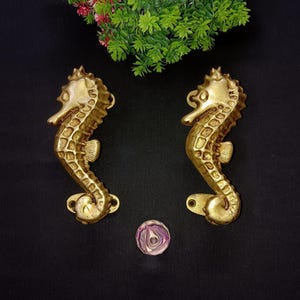 May include: A pair of gold-toned brass door handles shaped like seahorses. The handles have a detailed design and are mounted on a black background.
