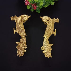 Puede incluir: Un par de tiradores de puerta de metal dorado con forma de pavo real con detalles intrincados. Los tiradores se enfrentan entre sí y tienen un diseño tradicional indio.