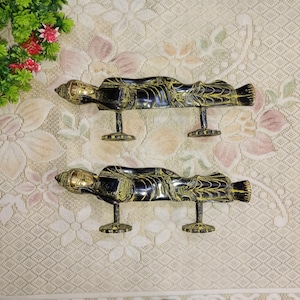 Op de afbeelding: Twee zwarte en gouden deurgrepen in de vorm van liggende Boeddha-figuren. De grepen zijn gemonteerd op een wit oppervlak met een bloemenpatroon.