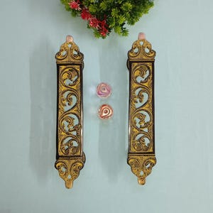 Może przedstawiać: Dwa ozdobne uchwyty do drzwi z misternymi wzorami z pozłacanego liścia na czarnym tle. Uchwyty są wykonane z drewna i mają tradycyjny styl.