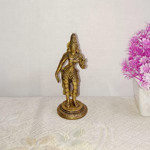 Puede incluir: Una estatua de latón de color dorado que representa a una diosa femenina con una corona y detalles intrincados. La estatua está de pie sobre una base redonda y sostiene un objeto pequeño en su mano derecha.