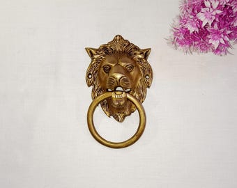 Puede incluir: Un llamador de puerta de cabeza de león dorado con un anillo de tracción. La cara del león está detallada con una melena y una boca cerrada.