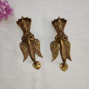 May include: Two gold-toned door handles shaped like mermaids with wings and a snake-like crown. The mermaids are in a prayer position with their hands clasped together.