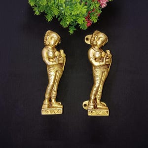 May include: A pair of gold-toned metal door handles shaped like women holding birds. The handles are detailed with intricate carvings and have a traditional Indian design.