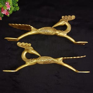 May include: Two gold-toned metal door handles shaped like peacocks with intricate details. The handles are in a horizontal position and have a curved shape.