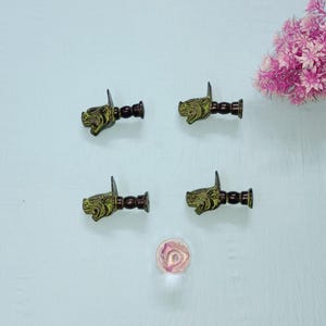 Op de afbeelding: Vier antieke messing haken in de vorm van een hondenkop met een donkerbruine steel en montageplaat. De hondenkoppen hebben een groen-gouden afwerking. Een klein, roze, rond glazen object is gecentreerd onder de haken. Een tros roze bloemen staat in de rechterbovenhoek.