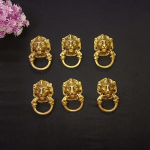 Puede incluir: Seis tiradores de cajón antiguos de latón con cabeza de león y asas de anillo. Las cabezas de león están detalladas con melenas y rasgos faciales. Los tiradores están dispuestos sobre un fondo oscuro, con flores rosas.