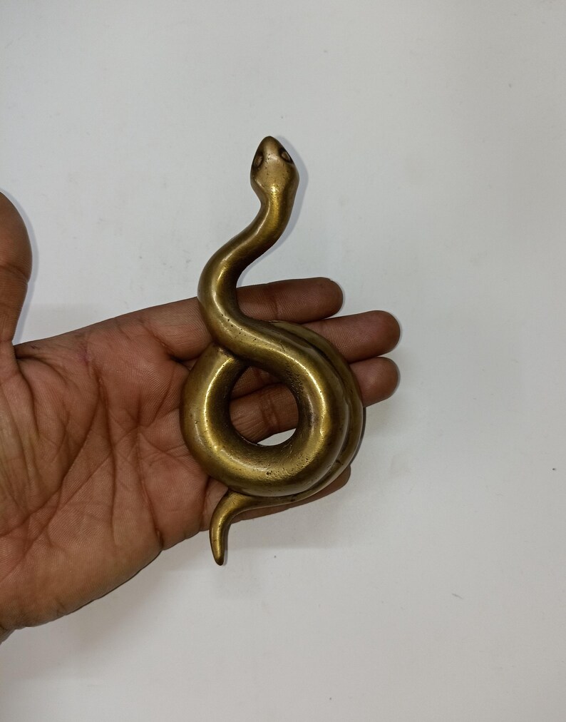 May include: A brass snake figurine with a coiled body and raised head. The sculpture has a smooth, polished finish and is a golden-brown colour. The snake is held in a hand against a white background.