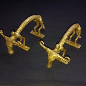 May include: Two gold-toned metal door handles shaped like giraffes with their heads bent down. The handles are mounted on rectangular metal plates.