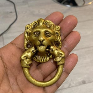 Op de afbeelding: Een gouden deurklopper in de vorm van een leeuwenkop met een ring die in de bek wordt gehouden door twee kleinere leeuwenkoppen. De klopper is gemaakt van metaal en heeft ingewikkelde details, waaronder een manen en gezichtskenmerken.