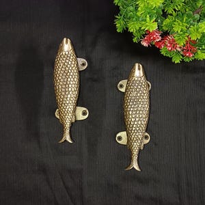 Peut inclure: Deux poignées de porte en forme de poisson dorées avec un design texturé et écailleux. Les poignées sont montées sur un fond noir.