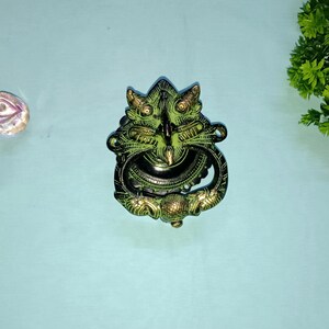 Op de afbeelding: Een groene en gouden metalen deurklopper in de vorm van een drakenkop met een ring om aan te trekken.